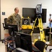 Three military personnel in OCP (Operational Camouflage Pattern) uniforms are gathered in what appears to be a technical laboratory or training room. One individual on the right appears to be briefing or demonstrating equipment to the other two. A Trimble surveying total station on a yellow tripod is prominently displayed in the foreground.