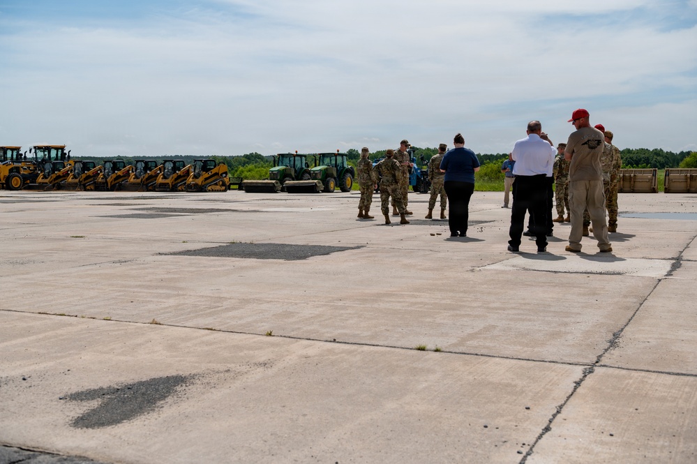 A group of military personnel in camouflage uniforms and several civilians are gathered on a large concrete surface, likely an airfield or military installation tarmac. A line of yellow construction equipment including skid steers, backhoes, and tractors is staged along the left side of the image. The scene appears to be a briefing, inspection, or pre-operation gathering related to construction or repair work.