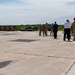 A group of military personnel in camouflage uniforms and several civilians are gathered on a large concrete surface, likely an airfield or military installation tarmac. A line of yellow construction equipment including skid steers, backhoes, and tractors is staged along the left side of the image. The scene appears to be a briefing, inspection, or pre-operation gathering related to construction or repair work.