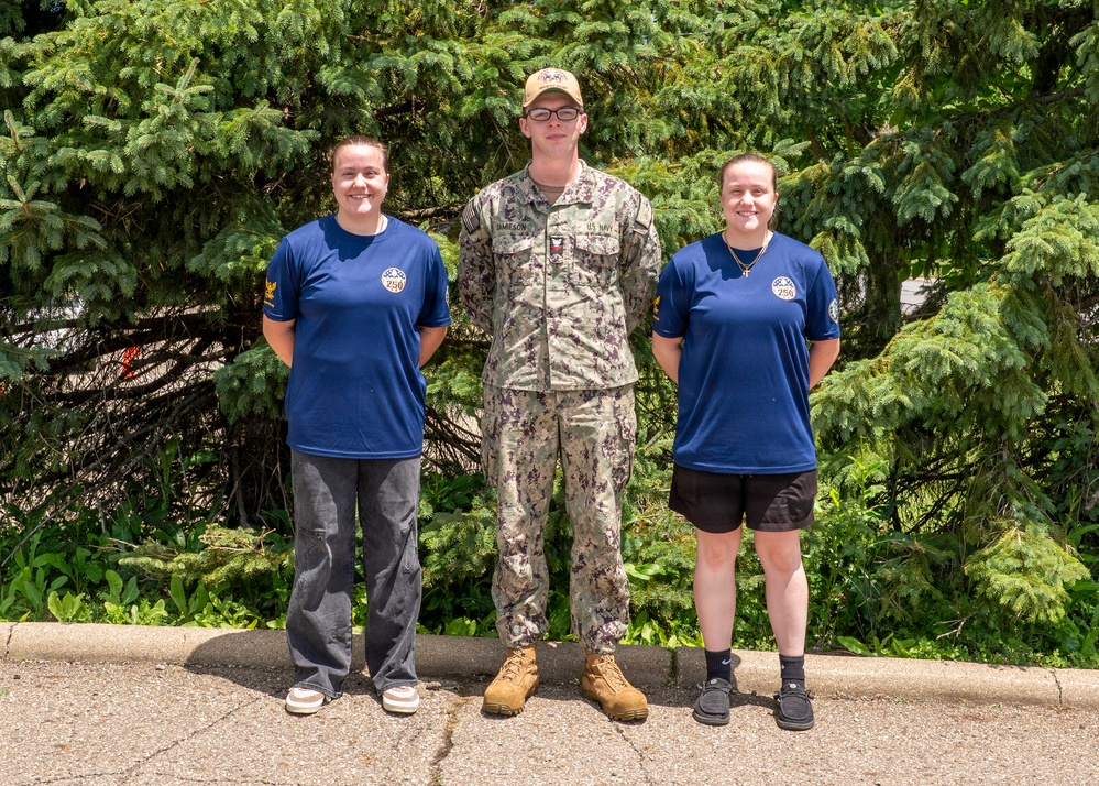 Three individuals pose for a group photo outdoors in front of evergreen trees. The center person is wearing a U.S. Navy NWU (Naval Working Uniform) camouflage uniform with a tan cap and name tape reading 'JAMESON', while the two flanking individuals wear matching blue t-shirts with what appears to be a '250' anniversary logo. The setting appears to be a paved outdoor area, likely at a military or naval installation.