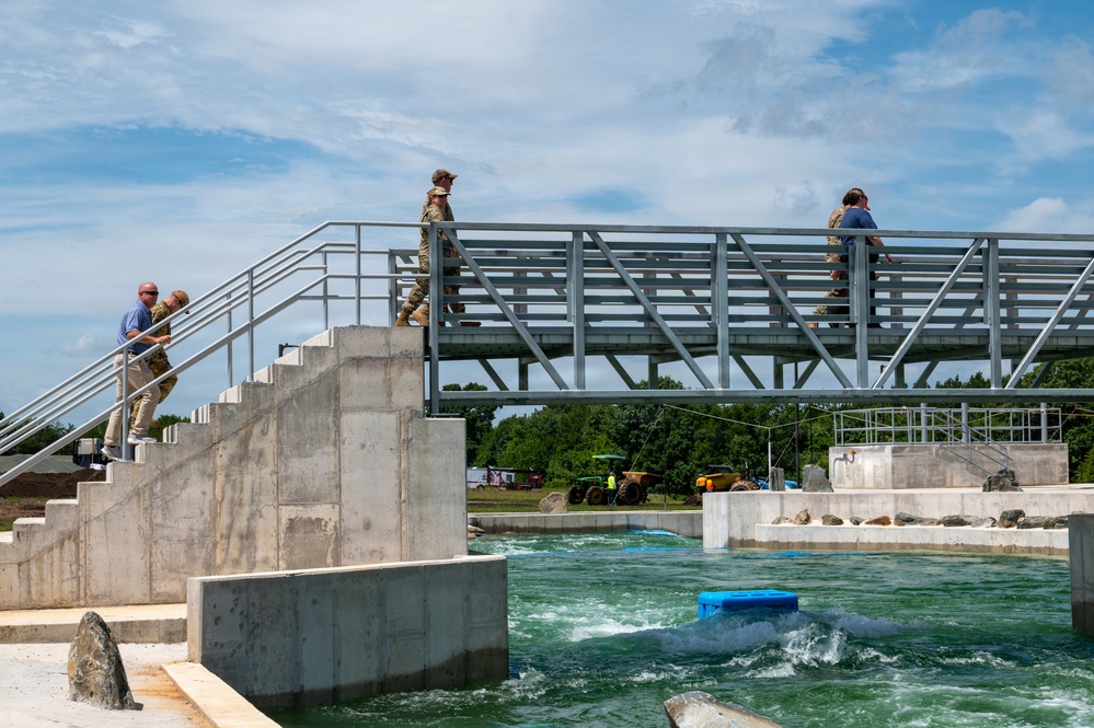 The image shows a group of individuals, some in military camouflage uniforms and others in civilian clothing, on and around a metal observation bridge overlooking an artificial whitewater channel or water feature facility. The structure includes concrete abutments, metal staircases, and railings, with turbulent green water flowing below. Construction equipment and vehicles are visible in the background, suggesting the facility may be newly built or still under development.