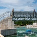 The image shows a group of individuals, some in military camouflage uniforms and others in civilian clothing, on and around a metal observation bridge overlooking an artificial whitewater channel or water feature facility. The structure includes concrete abutments, metal staircases, and railings, with turbulent green water flowing below. Construction equipment and vehicles are visible in the background, suggesting the facility may be newly built or still under development.
