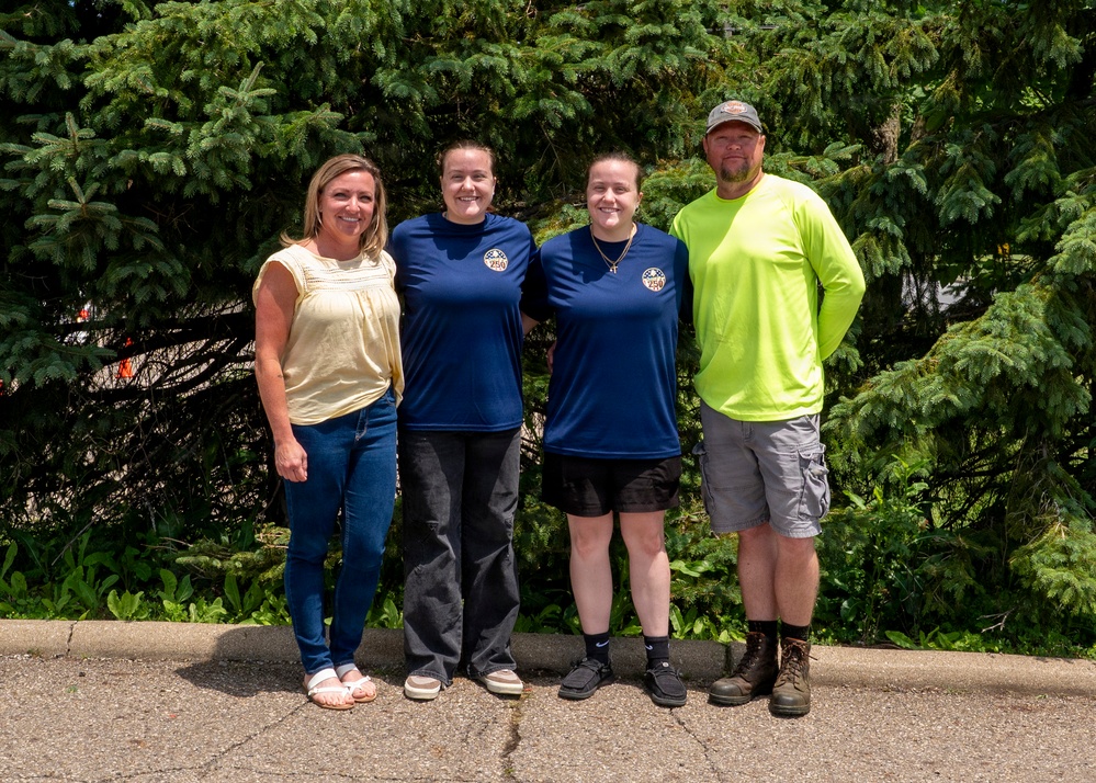 Four adults are posing together outdoors in front of a dense evergreen tree backdrop. Two of the individuals in the center are wearing matching navy blue shirts with a circular logo/patch. The group appears to be gathered for a casual group photo, likely at an outdoor event or workplace setting.