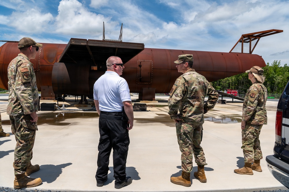 Four individuals, three in U.S. military camouflage uniforms (OCP pattern) and one civilian in a white polo shirt and black pants, are gathered outdoors examining or discussing a large rusted metal structure. The structure appears to be a large cylindrical tank or vessel mounted on a platform, showing significant oxidation and weathering. The setting appears to be a military installation or industrial facility on a clear, partly cloudy day.