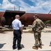Four individuals, three in U.S. military camouflage uniforms (OCP pattern) and one civilian in a white polo shirt and black pants, are gathered outdoors examining or discussing a large rusted metal structure. The structure appears to be a large cylindrical tank or vessel mounted on a platform, showing significant oxidation and weathering. The setting appears to be a military installation or industrial facility on a clear, partly cloudy day.