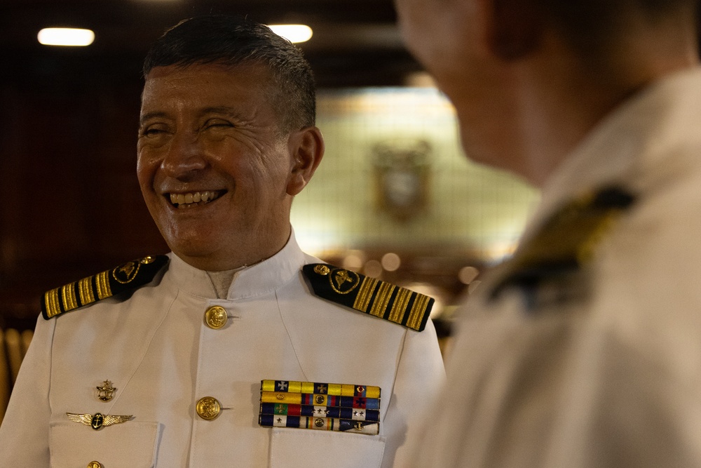 A senior naval officer in white dress uniform is smiling broadly while engaged in conversation with another uniformed individual whose back is partially visible in the foreground. The officer displays multiple service ribbons, qualification badges, and gold epaulettes indicating high rank. The setting appears to be an official naval facility or ceremonial space with warm ambient lighting.