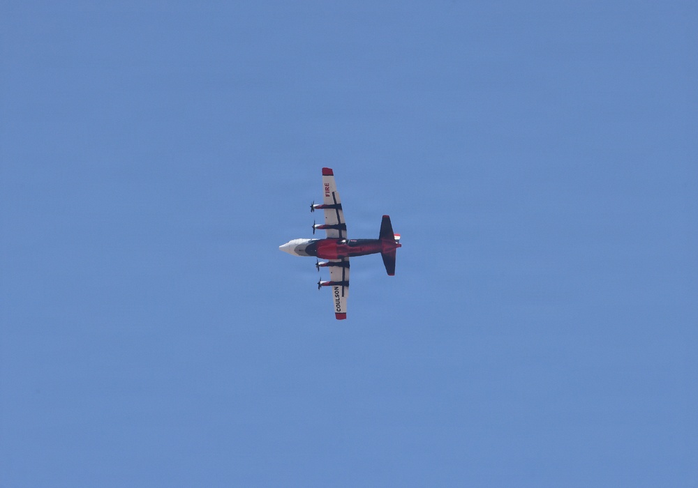 A firefighting aircraft is photographed from below against a clear blue sky. The plane is red and white with 'COULSON' visible on one wing and 'FIRE' on the tail, identifying it as a Coulson Aviation air tanker used for aerial firefighting operations. The image is taken from a ground-level perspective looking upward.