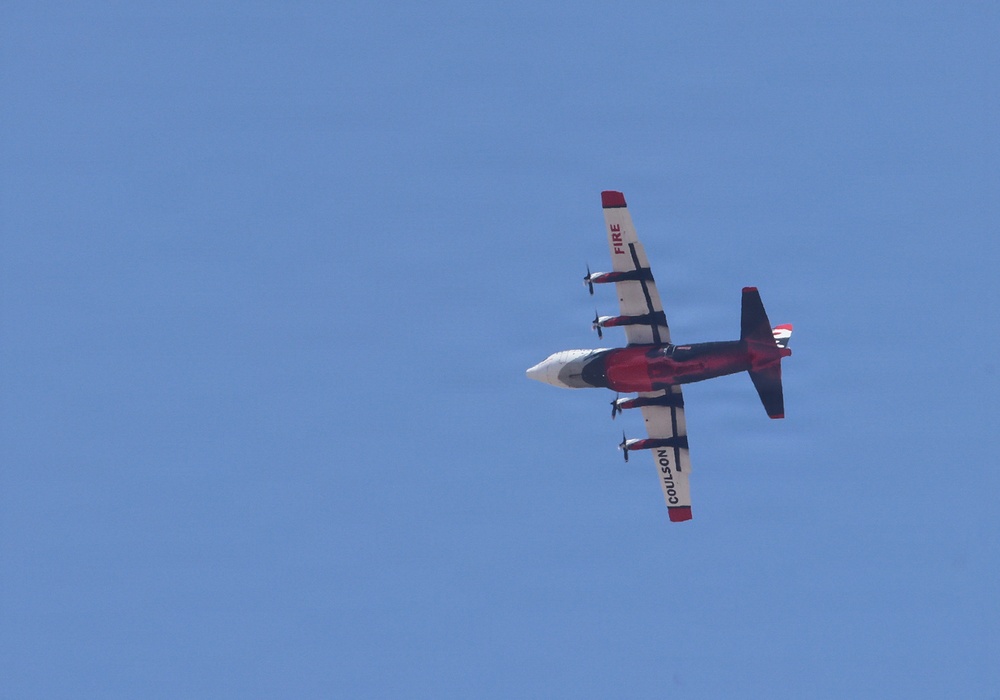 A red and white multi-engine turboprop aircraft is photographed from below against a clear blue sky. The aircraft bears the markings 'COULSON' on the wing and 'FIRE' on the tail, identifying it as a Coulson Aviation firefighting/aerial tanker aircraft. The plane appears to be in level flight, viewed from a ground-level perspective.
