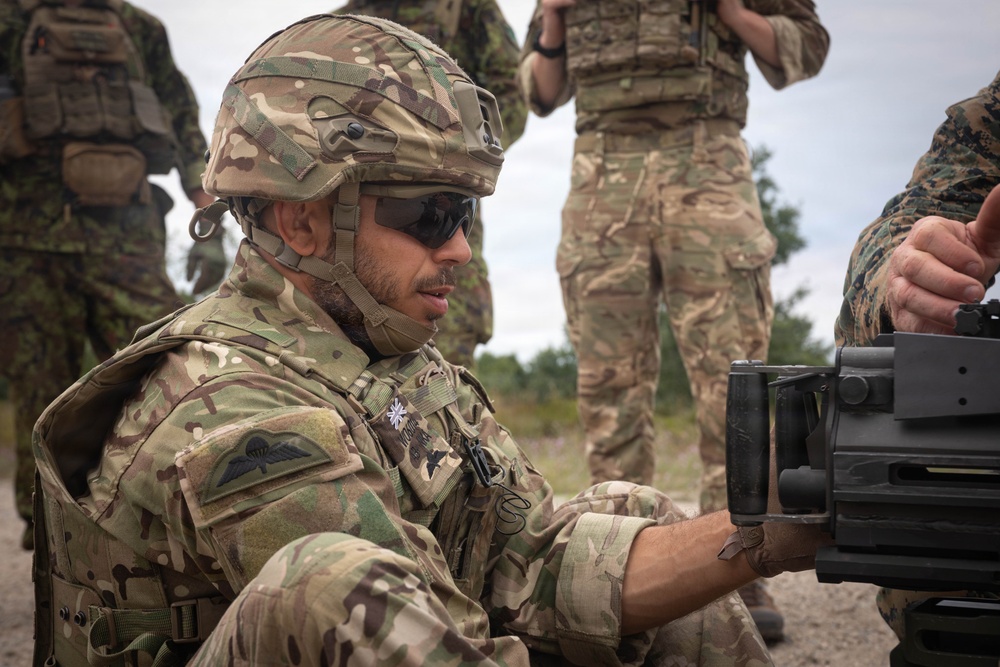 A soldier in MultiCam/MTP camouflage uniform, wearing a ballistic helmet and tactical sunglasses, is crouched down examining or handling a weapon system. Multiple other military personnel in various camouflage patterns are visible in the background. The setting appears to be an outdoor training or exercise environment.