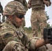 A soldier in MultiCam/MTP camouflage uniform, wearing a ballistic helmet and tactical sunglasses, is crouched down examining or handling a weapon system. Multiple other military personnel in various camouflage patterns are visible in the background. The setting appears to be an outdoor training or exercise environment.
