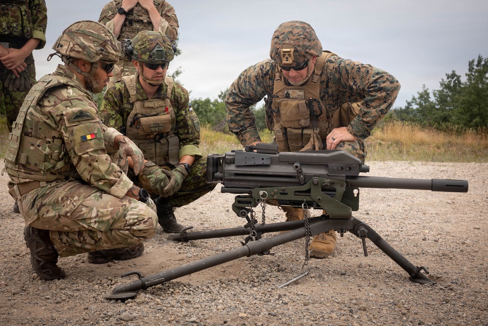 Three military personnel from different nations are gathered around a crew-served weapon system mounted on a tripod, appearing to conduct a familiarization or training exercise. One soldier displays a Romanian flag patch on their left shoulder, another wears what appears to be Canadian camouflage, and the third wears US Marine Corps MARPAT digital camouflage. The setting is an outdoor training range with gravel ground and vegetation in the background.