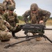 Three military personnel from different nations are gathered around a crew-served weapon system mounted on a tripod, appearing to conduct a familiarization or training exercise. One soldier displays a Romanian flag patch on their left shoulder, another wears what appears to be Canadian camouflage, and the third wears US Marine Corps MARPAT digital camouflage. The setting is an outdoor training range with gravel ground and vegetation in the background.