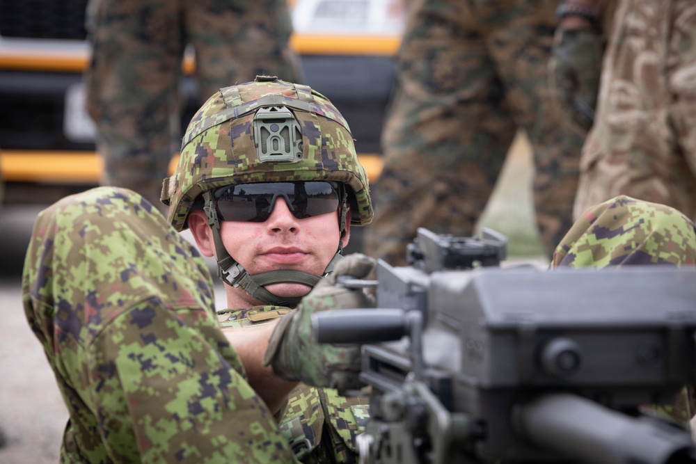 A soldier in digital camouflage uniform and helmet is positioned behind a crew-served weapon, looking directly at the camera. The soldier wears tactical sunglasses and a modern ballistic helmet with a mount attachment. Additional personnel in mixed camouflage patterns are visible in the blurred background.