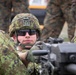 A soldier in digital camouflage uniform and helmet is positioned behind a crew-served weapon, looking directly at the camera. The soldier wears tactical sunglasses and a modern ballistic helmet with a mount attachment. Additional personnel in mixed camouflage patterns are visible in the blurred background.