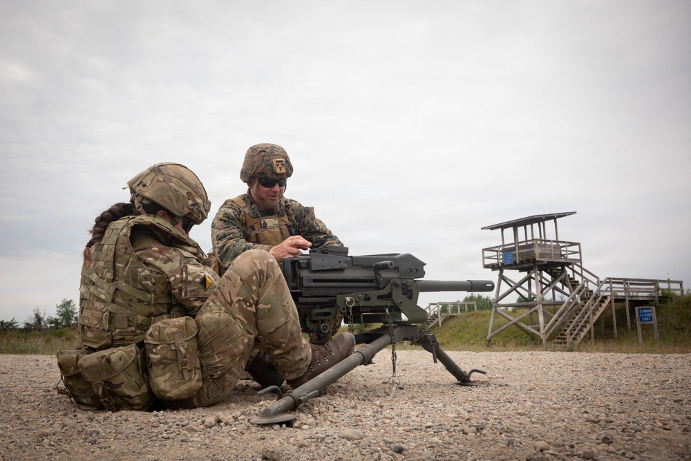 Two military personnel in camouflage uniforms are seated on a gravel surface at what appears to be a firing range, examining or operating a crew-served weapon system mounted on a tripod. One soldier appears to be instructing the other on the weapon's operation. A wooden observation tower is visible in the background.