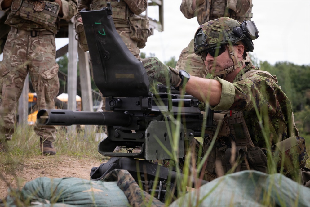 A soldier in camouflage uniform and tactical helmet is operating what appears to be an Mk 19 grenade launcher or similar crew-served weapon mounted on a tripod. The setting appears to be a military training range with other uniformed personnel visible in the background. The soldier is wearing full tactical gear including a combat helmet with night vision mount, sunglasses, and a wristwatch.