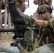 A soldier in camouflage uniform and tactical helmet is operating what appears to be an Mk 19 grenade launcher or similar crew-served weapon mounted on a tripod. The setting appears to be a military training range with other uniformed personnel visible in the background. The soldier is wearing full tactical gear including a combat helmet with night vision mount, sunglasses, and a wristwatch.