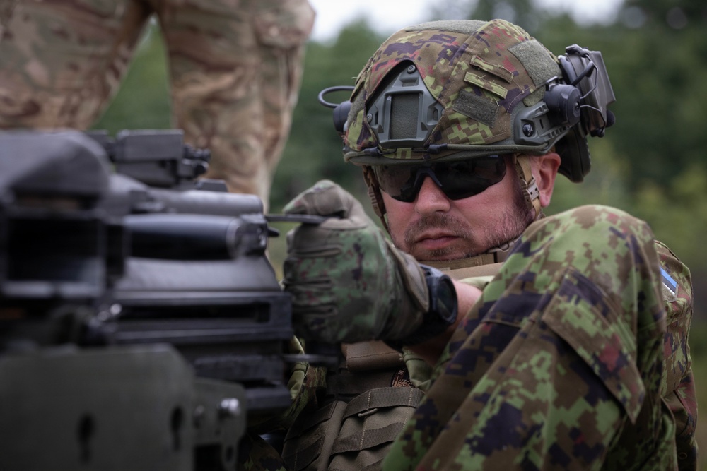 A soldier in modern combat gear is closely inspecting or operating a crew-served weapon system, likely a machine gun or grenade launcher. The soldier wears distinctive pixelated camouflage consistent with Estonian Defence Forces pattern, along with a tactical helmet, hearing protection, and ballistic sunglasses. A second soldier in a different camouflage pattern is partially visible in the background.