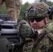 A soldier in modern combat gear is closely inspecting or operating a crew-served weapon system, likely a machine gun or grenade launcher. The soldier wears distinctive pixelated camouflage consistent with Estonian Defence Forces pattern, along with a tactical helmet, hearing protection, and ballistic sunglasses. A second soldier in a different camouflage pattern is partially visible in the background.