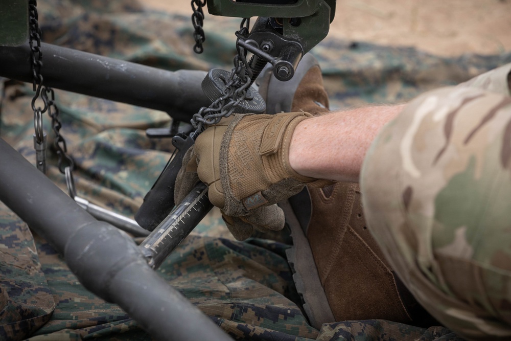 A military service member wearing MultiCam/OCP camouflage uniform and tan Mechanix tactical gloves is performing maintenance or assembly work on military equipment. The person is handling a graduated/scaled rod or tube component connected to a chain rigging system. The work is being conducted on a camouflage tarp laid over sandy ground.