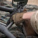 A military service member wearing MultiCam/OCP camouflage uniform and tan Mechanix tactical gloves is performing maintenance or assembly work on military equipment. The person is handling a graduated/scaled rod or tube component connected to a chain rigging system. The work is being conducted on a camouflage tarp laid over sandy ground.