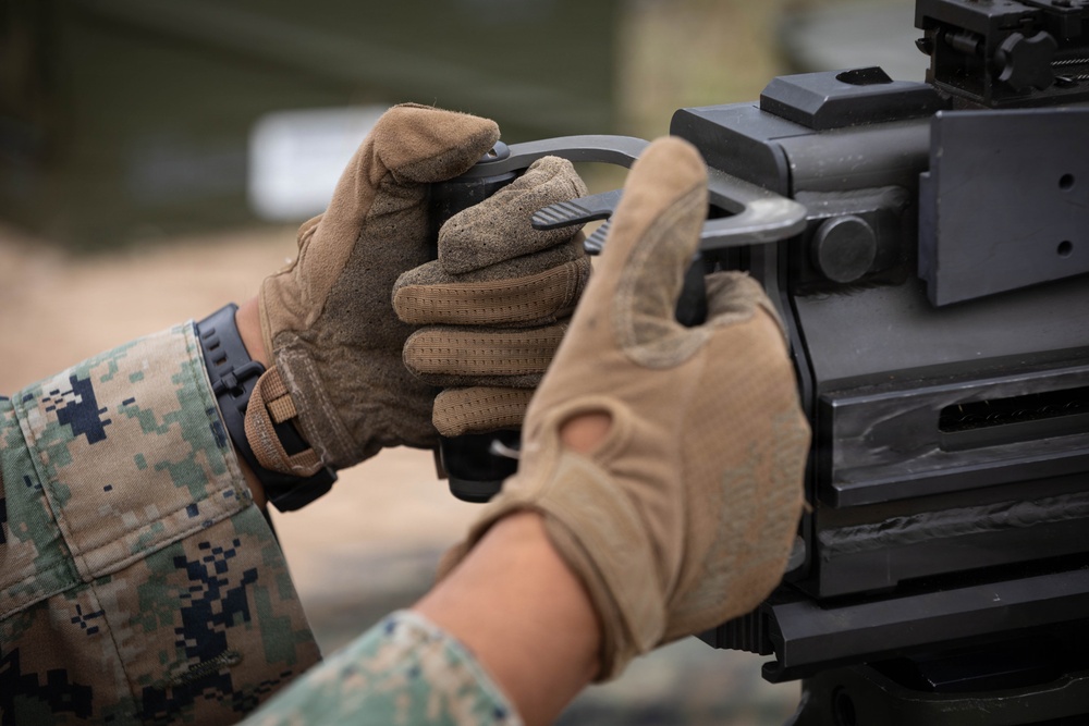 A close-up image showing the gloved hands of a military service member handling a heavy weapon system. The individual is wearing MARPAT (Marine Pattern) digital camouflage uniform, indicating U.S. Marine Corps affiliation. The focus is on the dual-grip handle area of what appears to be a crew-served machine gun or grenade launcher.