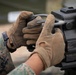 A close-up image showing the gloved hands of a military service member handling a heavy weapon system. The individual is wearing MARPAT (Marine Pattern) digital camouflage uniform, indicating U.S. Marine Corps affiliation. The focus is on the dual-grip handle area of what appears to be a crew-served machine gun or grenade launcher.