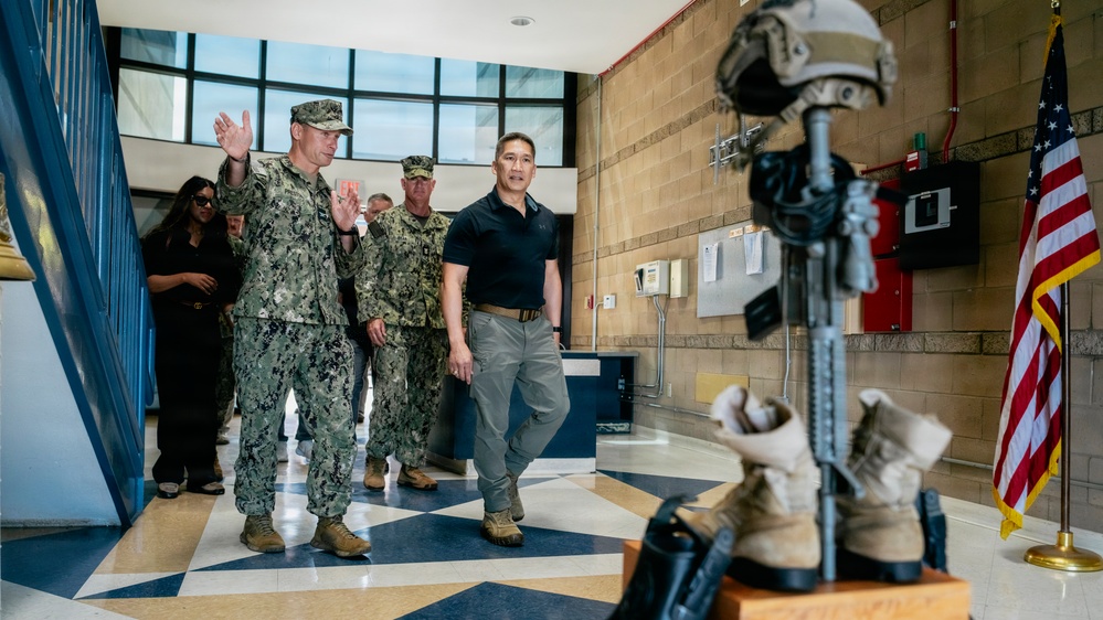 The image shows military personnel in Navy camouflage uniforms walking through what appears to be a military facility lobby alongside a civilian in a black polo shirt and khaki pants. In the foreground, a battlefield cross memorial display is visible, consisting of a rifle, boots, helmet, and gloves mounted on a wooden base, with an American flag nearby. The group appears to be engaged in a briefing or facility tour.