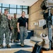 The image shows military personnel in Navy camouflage uniforms walking through what appears to be a military facility lobby alongside a civilian in a black polo shirt and khaki pants. In the foreground, a battlefield cross memorial display is visible, consisting of a rifle, boots, helmet, and gloves mounted on a wooden base, with an American flag nearby. The group appears to be engaged in a briefing or facility tour.