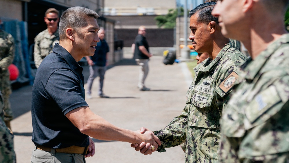 A civilian in a dark polo shirt is shaking hands with a U.S. Navy sailor in NWU Type III digital camouflage uniform during what appears to be an official visit or greeting ceremony. Additional military personnel in similar Navy uniforms are lined up, suggesting a formal reception or inspection. The setting appears to be an outdoor military installation or base.