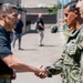 A civilian in a dark polo shirt is shaking hands with a U.S. Navy sailor in NWU Type III digital camouflage uniform during what appears to be an official visit or greeting ceremony. Additional military personnel in similar Navy uniforms are lined up, suggesting a formal reception or inspection. The setting appears to be an outdoor military installation or base.