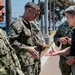 A group of U.S. Navy personnel in NWU Type III camouflage uniforms are gathered outdoors near a waterfront, appearing to be at a naval installation. A civilian in a dark polo shirt is holding a small red item in his outstretched palm, which is being examined by uniformed personnel. The setting appears to be San Diego, California, based on the visible bridge and palm trees in the background.