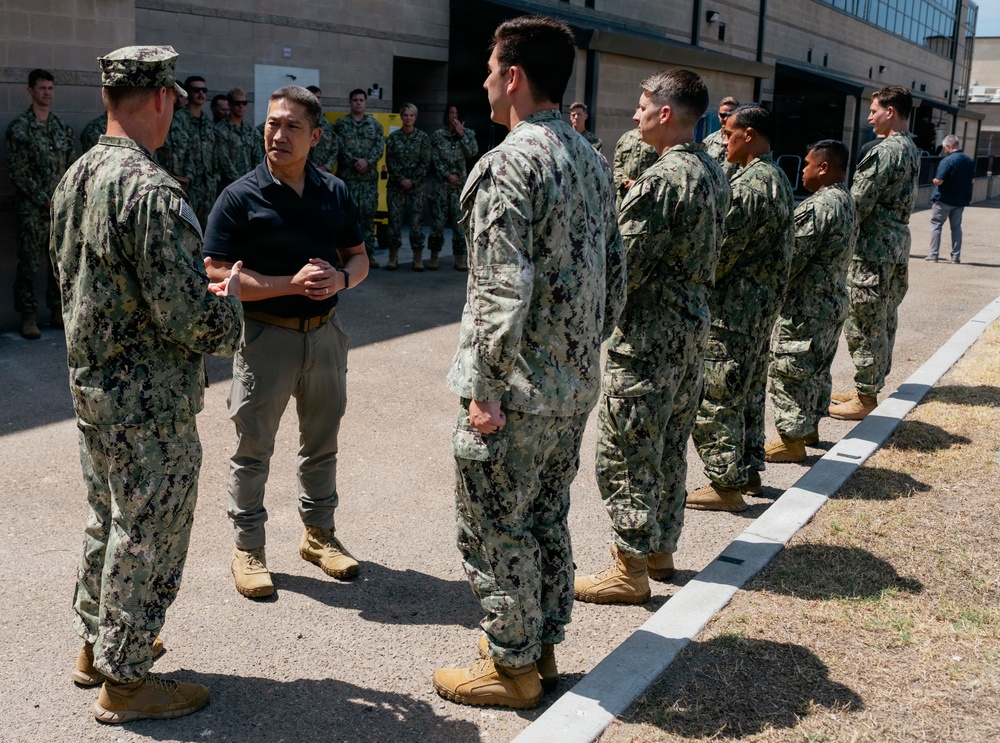 A civilian official in a black polo shirt and khaki pants is engaged in conversation with a uniformed military officer wearing Navy Working Uniform (NWU Type III camouflage) and a cover. A formation of Navy personnel in matching camouflage uniforms and tan boots stands at attention in a line, suggesting an official visit, inspection, or greeting ceremony at a military installation.