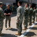 A civilian official in a black polo shirt and khaki pants is engaged in conversation with a uniformed military officer wearing Navy Working Uniform (NWU Type III camouflage) and a cover. A formation of Navy personnel in matching camouflage uniforms and tan boots stands at attention in a line, suggesting an official visit, inspection, or greeting ceremony at a military installation.