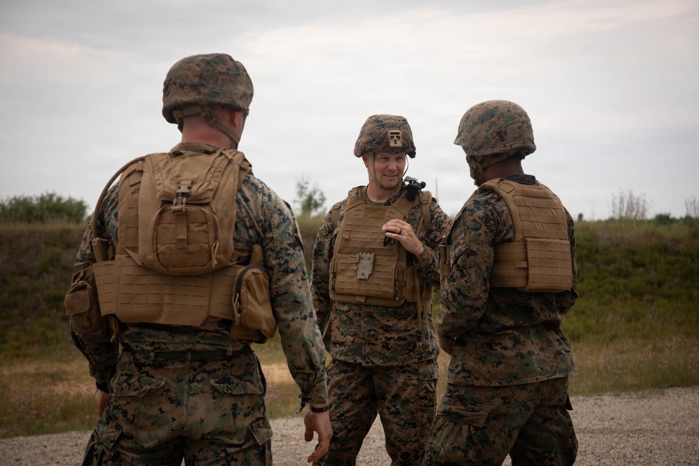 Three military personnel in MARPAT (Marine pattern) camouflage uniforms are engaged in a conversation in an outdoor field environment. All three are wearing tactical gear including plate carriers and helmets, with one individual appearing to hold a radio or communication device. The setting appears to be a training area or operational environment with vegetation and a dirt path visible in the background.