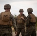 Three military personnel in MARPAT (Marine pattern) camouflage uniforms are engaged in a conversation in an outdoor field environment. All three are wearing tactical gear including plate carriers and helmets, with one individual appearing to hold a radio or communication device. The setting appears to be a training area or operational environment with vegetation and a dirt path visible in the background.