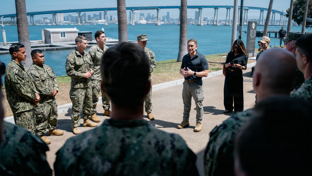 A civilian man in a black polo shirt and khaki pants addresses a group of military personnel in camouflage uniforms outdoors near a waterfront. The setting appears to be a naval installation with a large bridge and city skyline visible in the background across the bay. The gathering has the appearance of a briefing or informal address to service members.