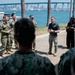 A civilian man in a black polo shirt and khaki pants addresses a group of military personnel in camouflage uniforms outdoors near a waterfront. The setting appears to be a naval installation with a large bridge and city skyline visible in the background across the bay. The gathering has the appearance of a briefing or informal address to service members.