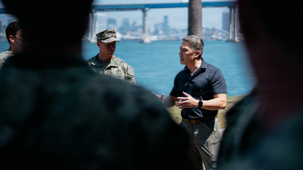 A civilian in a dark polo shirt appears to be briefing or addressing a group that includes at least one military personnel in camouflage uniform. The setting is outdoors near a waterfront with a large bridge and city skyline visible in the background, suggesting a coastal urban location. The scene has the appearance of an official or professional outdoor meeting.