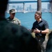 A civilian in a dark polo shirt appears to be briefing or addressing a group that includes at least one military personnel in camouflage uniform. The setting is outdoors near a waterfront with a large bridge and city skyline visible in the background, suggesting a coastal urban location. The scene has the appearance of an official or professional outdoor meeting.
