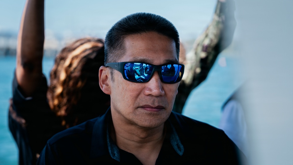 A middle-aged man wearing blue mirrored wraparound sunglasses and a black shirt is the primary subject, photographed in close-up with a shallow depth of field. The background shows a blurred waterfront or harbor setting with other people partially visible. The reflections in his sunglasses suggest an indoor or sheltered environment nearby.