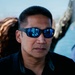 A middle-aged man wearing blue mirrored wraparound sunglasses and a black shirt is the primary subject, photographed in close-up with a shallow depth of field. The background shows a blurred waterfront or harbor setting with other people partially visible. The reflections in his sunglasses suggest an indoor or sheltered environment nearby.