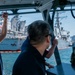 A man wearing a dark polo shirt and glasses is viewed from behind aboard a small vessel, observing naval warships docked in a harbor. At least two large military ships are visible in the background, with partial text readable on one hull appearing to read 'FITZGERALD'. Multiple people are present on the observation vessel, suggesting a tour or official visit to a naval installation.