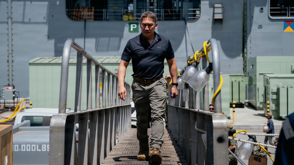 A man in civilian attire walks down a gangway or ramp from a large naval vessel at a military port or dock facility. He is dressed in a dark polo shirt and tactical-style pants with boots, suggesting a professional or official visit. The setting appears to be an active naval installation with personnel and equipment visible in the background.