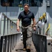 A man in civilian attire walks down a gangway or ramp from a large naval vessel at a military port or dock facility. He is dressed in a dark polo shirt and tactical-style pants with boots, suggesting a professional or official visit. The setting appears to be an active naval installation with personnel and equipment visible in the background.