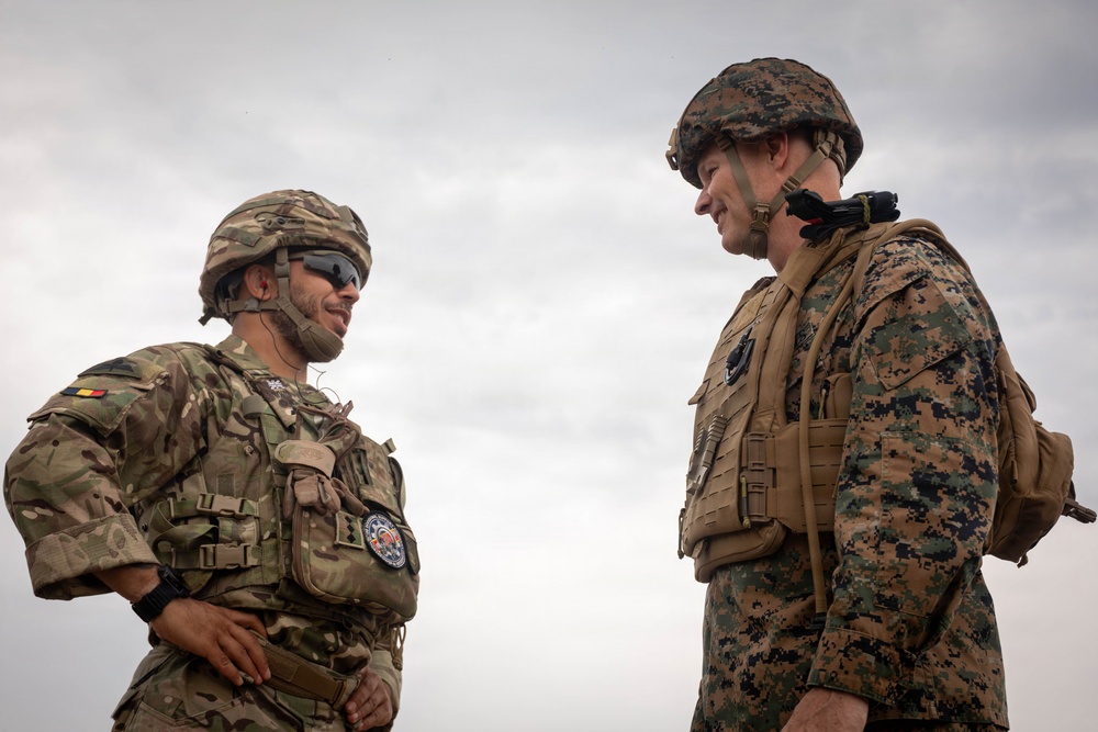 Two military personnel in full combat gear are engaged in a face-to-face conversation outdoors under an overcast sky. The soldier on the left wears MultiCam/OCP pattern camouflage with what appears to be a Romanian flag patch, while the soldier on the right wears MARPAT (Marine pattern) digital camouflage consistent with U.S. Marine Corps uniform. Both are equipped with tactical vests, helmets, and appear relaxed, suggesting a friendly or professional exchange.