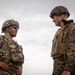Two military personnel in full combat gear are engaged in a face-to-face conversation outdoors under an overcast sky. The soldier on the left wears MultiCam/OCP pattern camouflage with what appears to be a Romanian flag patch, while the soldier on the right wears MARPAT (Marine pattern) digital camouflage consistent with U.S. Marine Corps uniform. Both are equipped with tactical vests, helmets, and appear relaxed, suggesting a friendly or professional exchange.