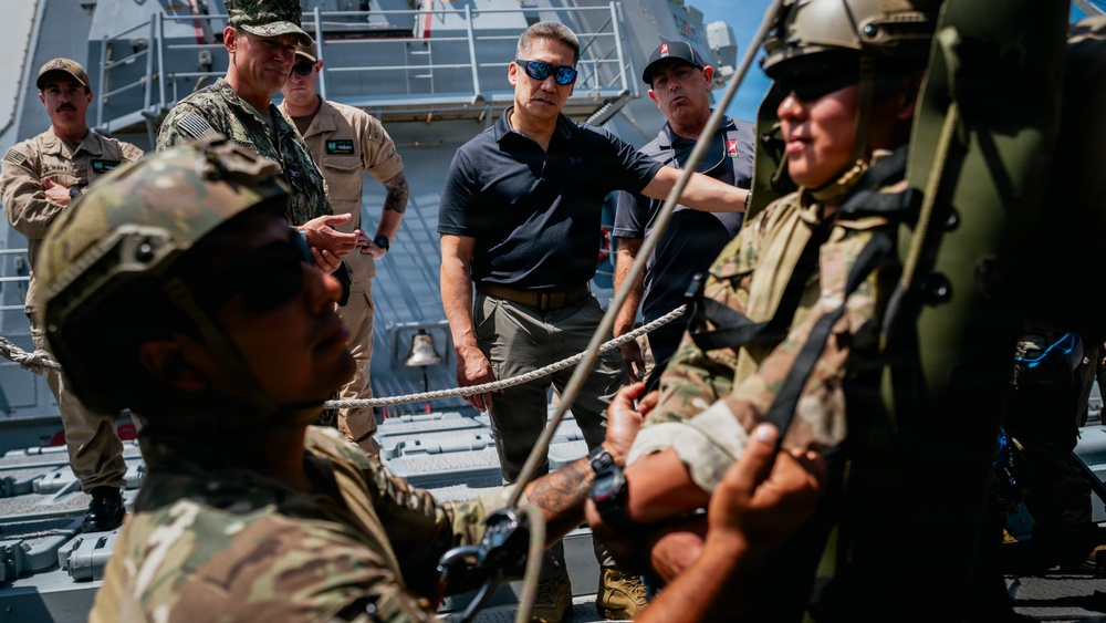 A group of military personnel and civilians are gathered on the deck of a naval vessel, observing or participating in what appears to be a rope/rigging demonstration or training exercise. Several individuals are in military camouflage uniforms with tactical gear, while others are in civilian or naval attire. The setting is clearly aboard a warship, with naval superstructure visible in the background.