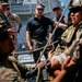 A group of military personnel and civilians are gathered on the deck of a naval vessel, observing or participating in what appears to be a rope/rigging demonstration or training exercise. Several individuals are in military camouflage uniforms with tactical gear, while others are in civilian or naval attire. The setting is clearly aboard a warship, with naval superstructure visible in the background.