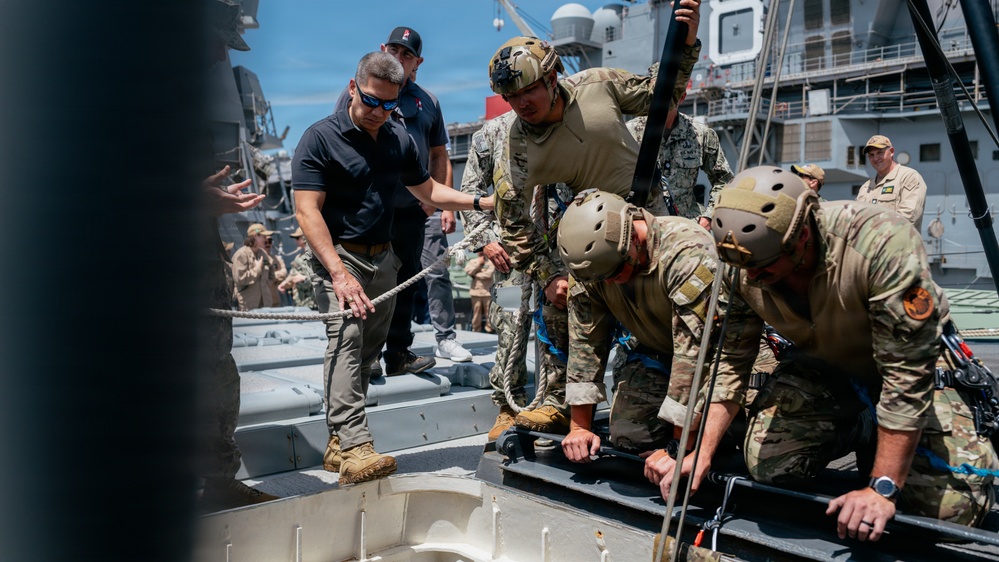 Military and civilian personnel are gathered on the deck of a naval vessel, working with ropes and rigging equipment. Several individuals wearing tactical helmets and camouflage uniforms appear to be conducting a training exercise or operational preparation involving rope systems. A civilian in a black polo shirt and sunglasses is actively participating alongside uniformed military members.